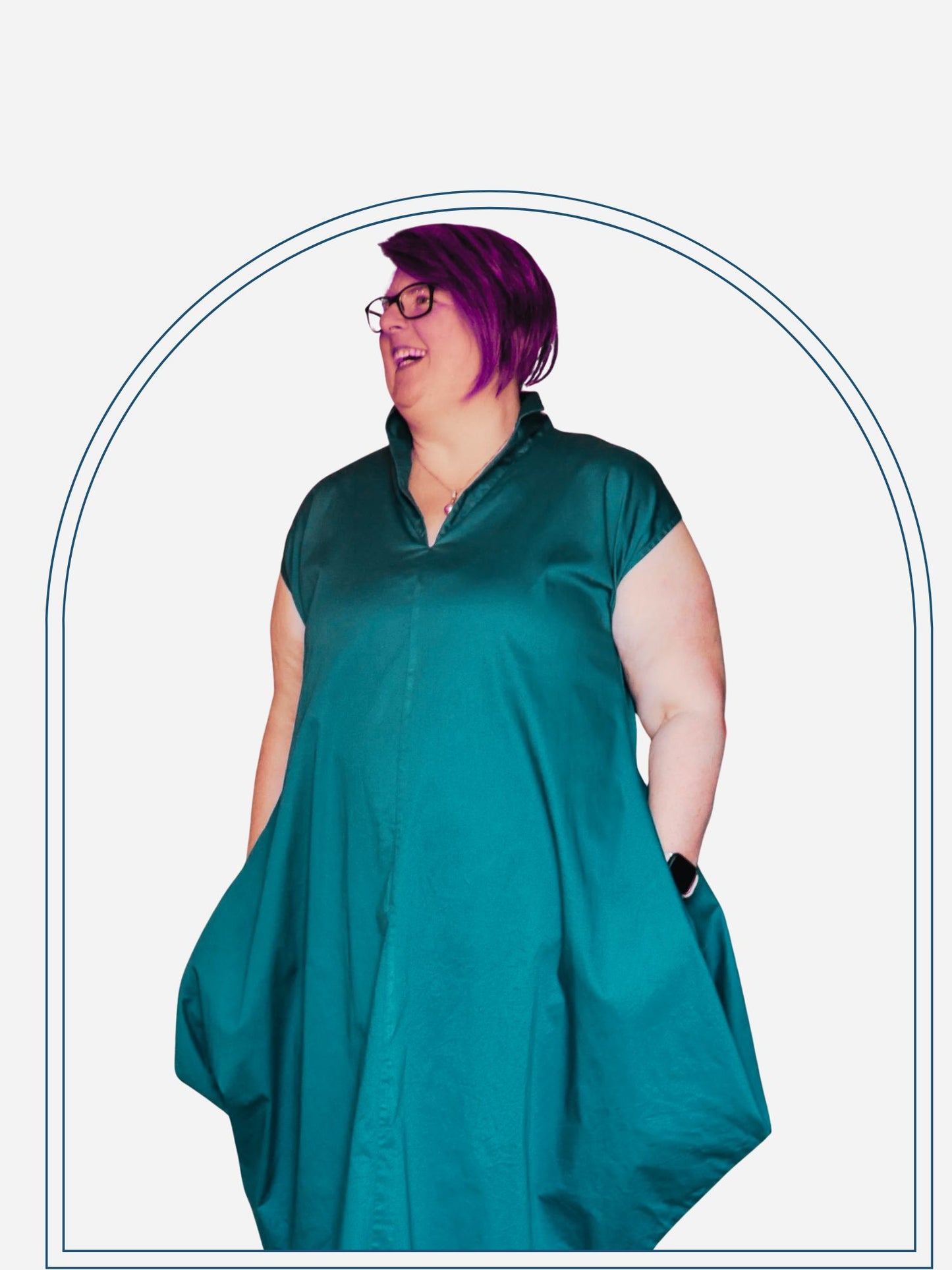 A woman standing in a home environment wearing a blue dress with a round neckline and long sleeves. The dress has a loose, draping fit and features pockets on the hips.