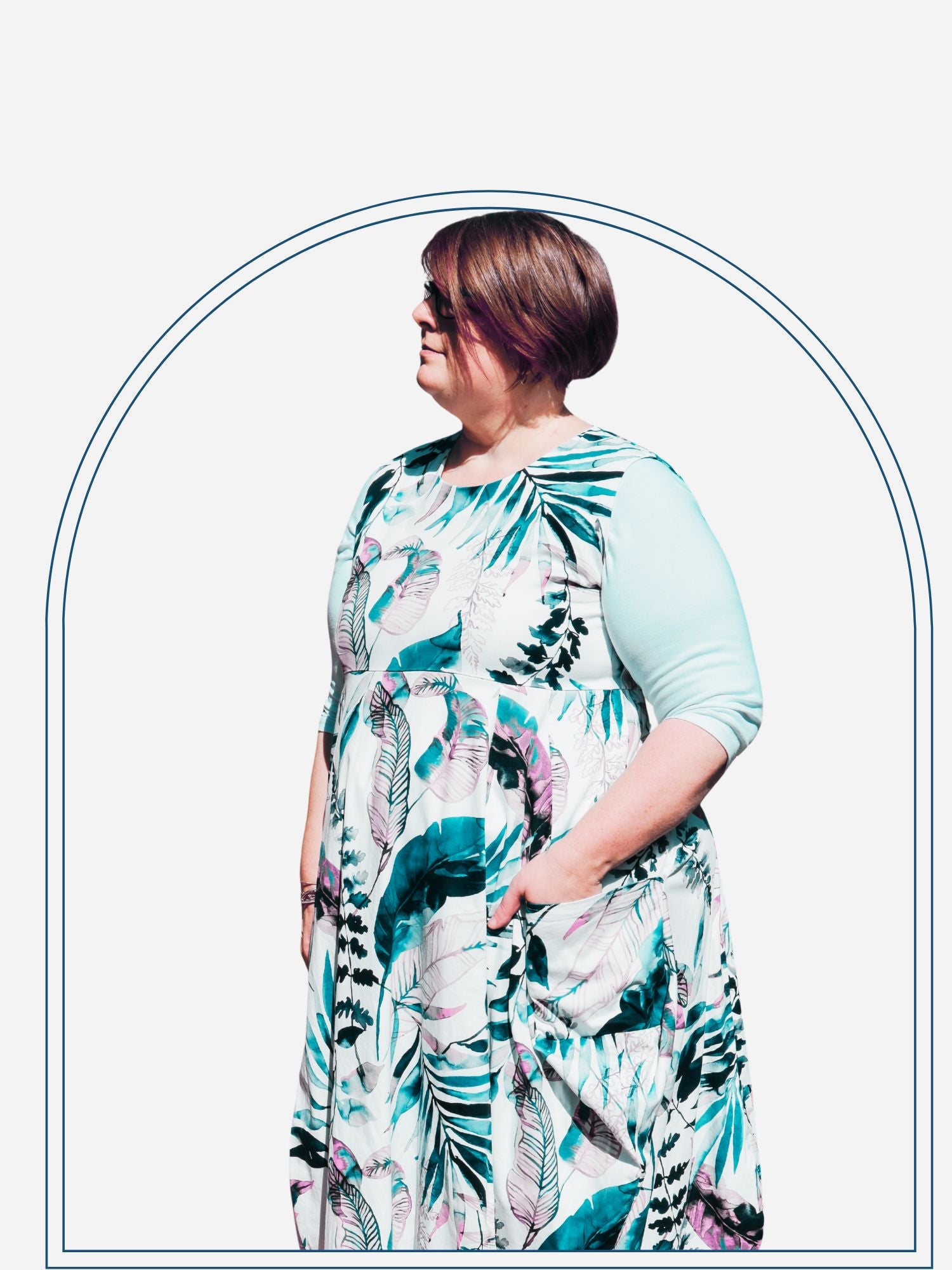 A woman wearing a floral print knee-length dress with sleeves, standing in front of a shrub.