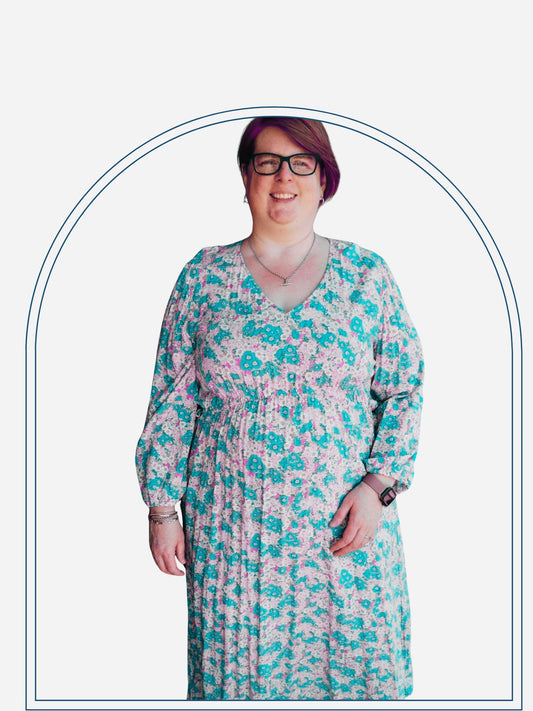 A woman standing on a deck wearing a flowing mid-length blue and white floral dress with 3/4 elasticated sleeves, waist ties, and an arched waistline, smiling at the camera.