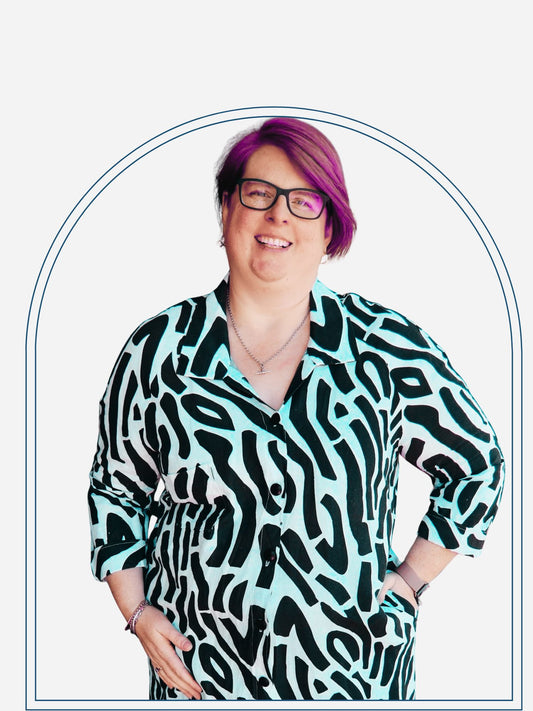 A woman standing on a deck wearing a longline shirt dress with a black and white zebra pattern, raglan sleeves, a collar, and buttons. She is also wearing glasses and a necklace.