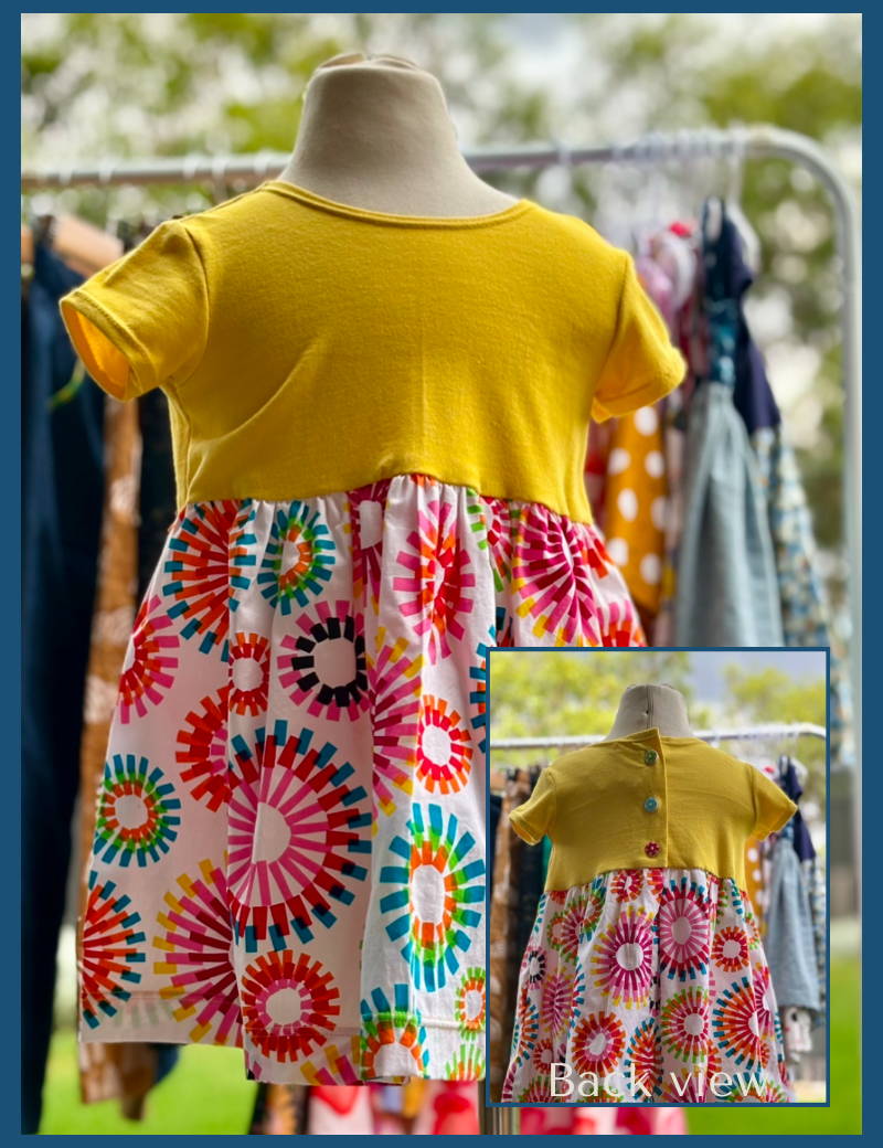 A yellow baby dress with a multicolored geometric pattern on the skirt, displayed on a mannequin, with a close-up of the back buttons.
