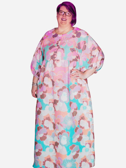 A woman standing in a colorful floral sleeve dress with pockets, posing against a modern art background.