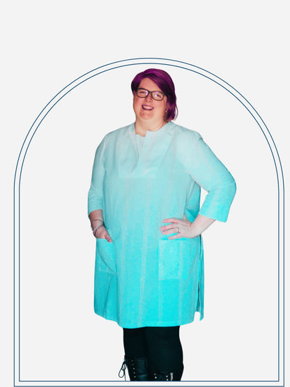 A woman standing in a doorway wearing a white tunic dress with 3/4 sleeves, paired with black boots and glasses.