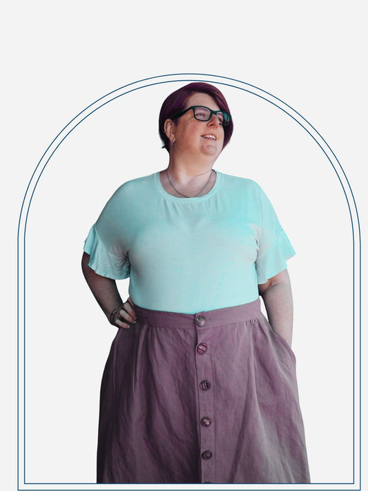 A woman standing outdoors wearing a sage green button-through skirt with natural wooden buttons, paired with a white top and glasses.